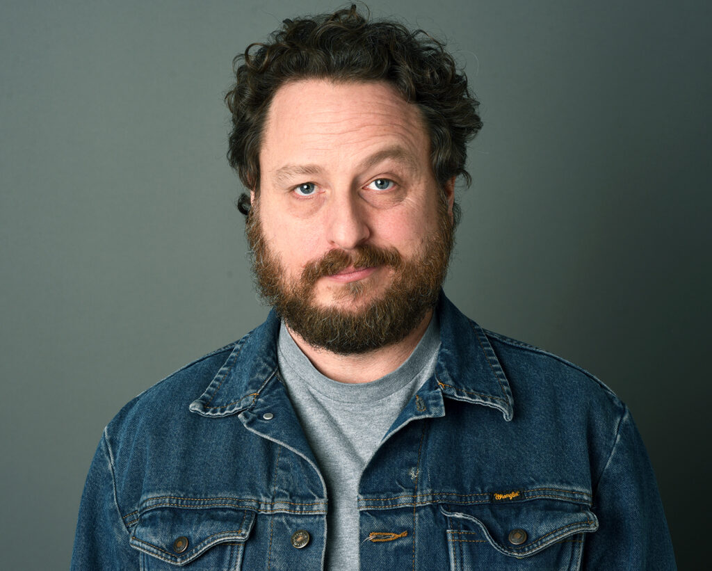 Gregg Brown, artist and actor, smirking at the camera, wearing a trendy jean jacket and a casual gray t-shirt.